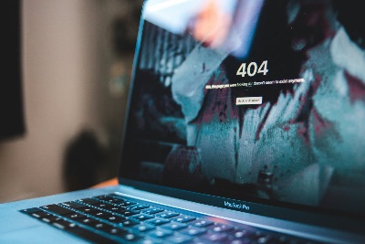Auf dem Bild ist ein geöffneter MacBook Pro Laptop zu sehen, dessen Bildschirm eine 404-Fehlerseite anzeigt. Die Fehlermeldung lautet "404 - Hmm, the page you were looking for doesn't seem to exist anymore" mit einem Button "Back to Unsplash". Das Bild hat eine künstlerische, leicht unscharfe Ästhetik mit bläulichen Farbtönen. Man erkennt die Tastatur des MacBook Pro im Vordergrund, während der Bildschirm im Fokus steht und die 404-Fehlermeldung deutlich sichtbar ist. Der Hintergrund ist bewusst unscharf gehalten, was den Fokus auf den Laptop und die Fehlermeldung lenkt. Es handelt sich um eine stilisierte Darstellung eines typischen Internet-Phänomens - der "Seite nicht gefunden"-Meldung, die jeder Internetnutzer kennt.